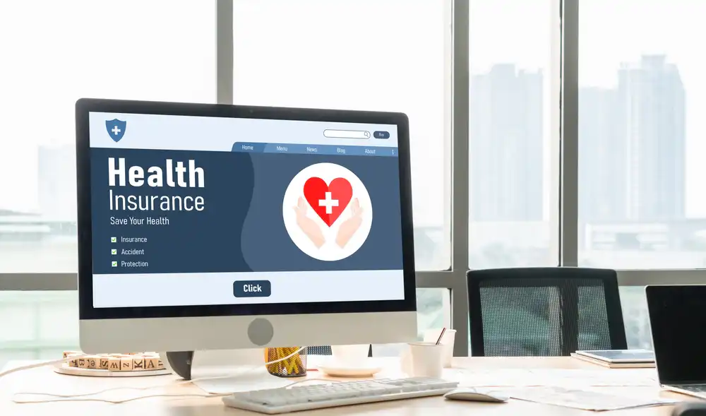 Desktop computer showing a health insurance website with a heart icon and 'Health Insurance' headline on a bright office desk.