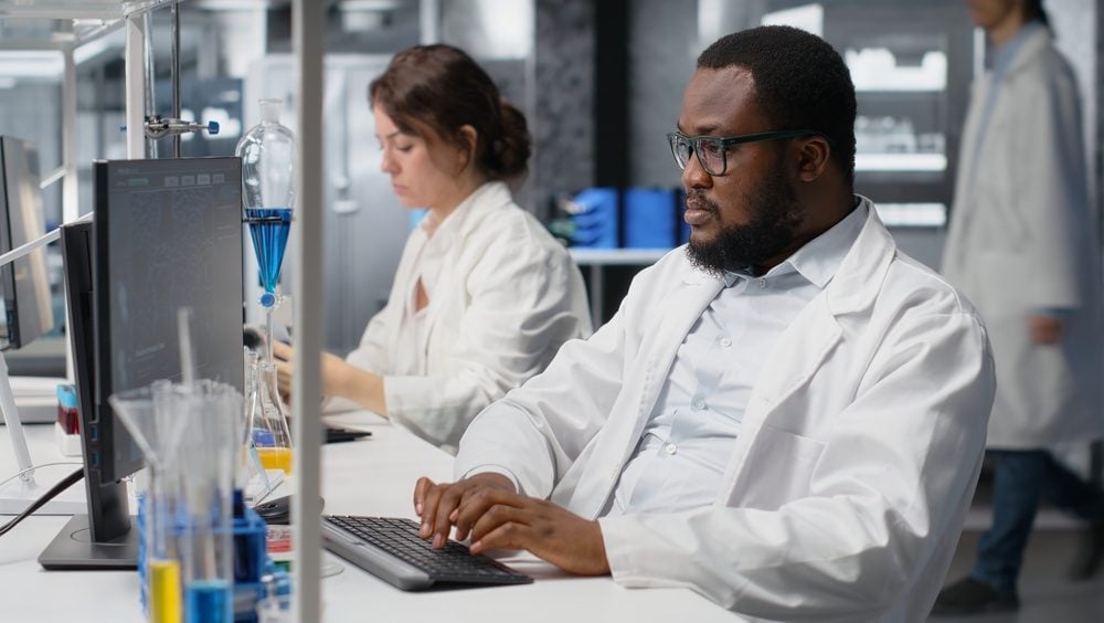 Two scientists in white lab coats at a bench, reviewing data on monitors near test tubes