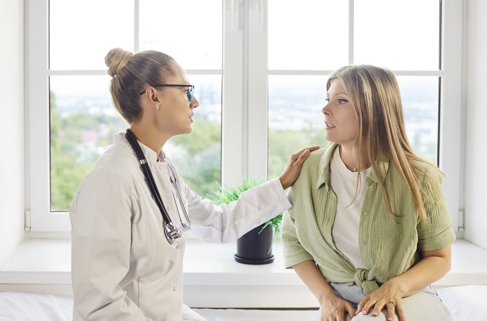 A doctor places a hand on a patient's shoulder while they talk together near a window.