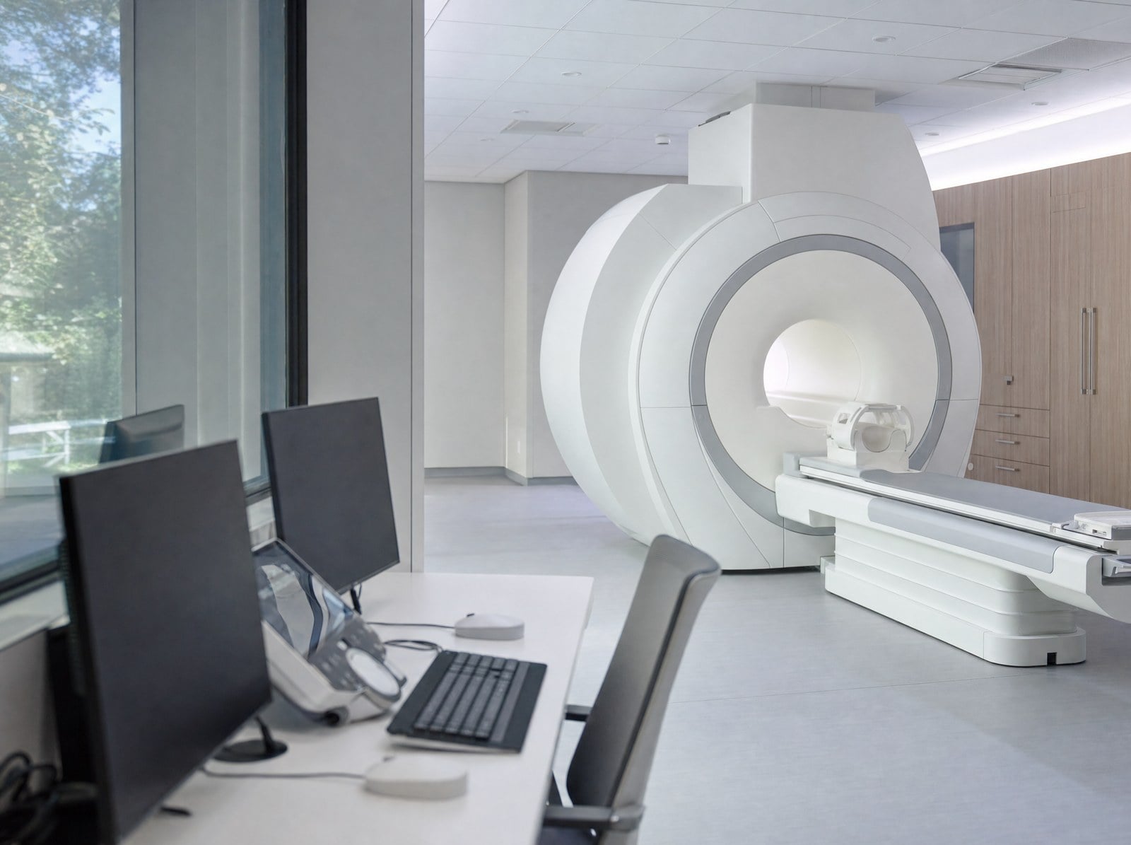 Two computer monitors at an MRI control desk, with the scanner bore visible in the room beyond.